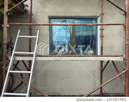 House facade under renovation with rusty scaffolding, aluminum ladder, covered window with protective plastic film and freshly plastered exterior wall, representing home improvement, construction work 141567765