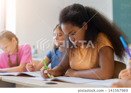 Focused black schoolgirl writing carefully in open notebook during classroom assignment Focused black schoolgirl writing carefully in open notebook during classroom assignment 141567969
