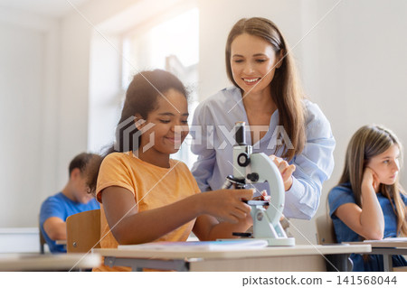 Smiling female teacher guiding black schoolgirl using microscope during practical science lesson Smiling female teacher guiding black schoolgirl using microscope during practical science lesson 141568044