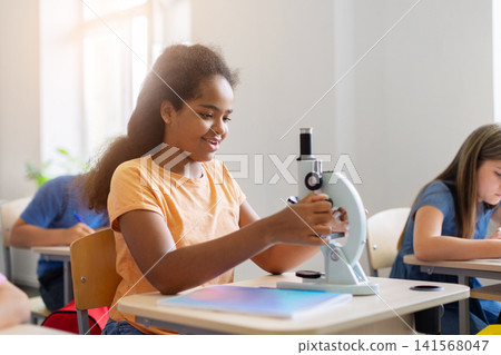 Smiling black schoolgirl adjusting microscope during engaging science lesson at her desk 141568047