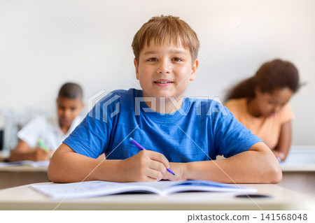 Smiling schoolboy sitting at classroom desk with an open workbook and pen 141568048