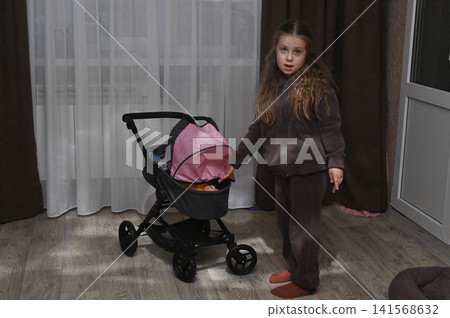 Little girl stands near toy baby stroller in room. Little girl stands near toy baby stroller in room. 141568632