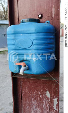 Blue plastic washbasin hangs on red wall for outdoor hand washing. 141568808