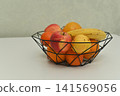 geometric wire basket filled with a mix of fruits like bananas, apples, and oranges. The fresh fruit appears vibrant against the plain white background, creating a clean and modern aesthetic. 141569056