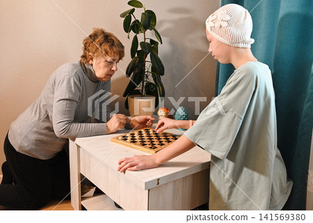 Elder woman and young girl playing checkers in a simple room with blue curtains Elder woman and young girl playing checkers in a simple room with blue curtains 141569380