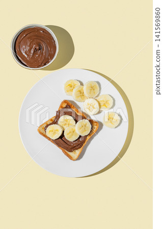 Toasted bread with chocolate cream and banana on a white plate on a yellow background Toasted bread with chocolate cream and banana on a white plate on a yellow background 141569860