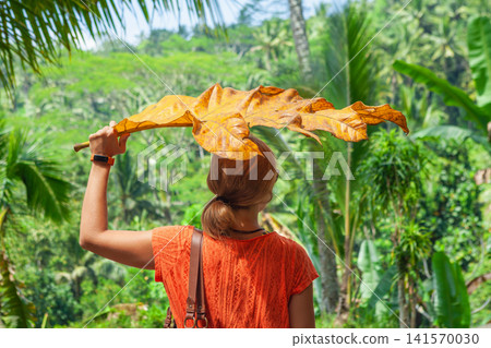 Back view of young woman under giant yellow tropical leaf while exploring jungle in Bali, Indonesia. Girl have a fun at family travel adventure on summer holidays. 141570030