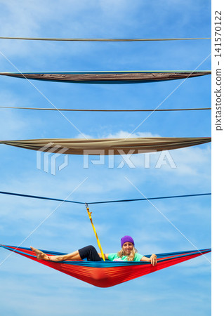 Young woman relaxing in hammock high above ground. A unique high-altitude attraction in Indonesia offering thrilling experiences and panoramic tropical forest views for adventure. Travel on vacation. Young woman relaxing in hammock high above ground. A unique high-altitude attraction in Indonesia offering thrilling experiences and panoramic tropical forest views for adventure. Travel on vacation. 141570122