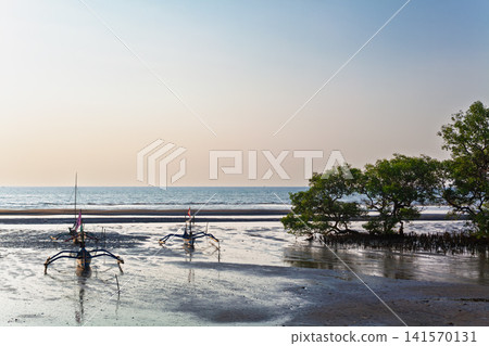 Traditional fishing boats rest on wet sand during low tide near mangroves. Calm sea, clear sky, warm tropical light create coastal scene perfect for travel, nature, and Indonesia lifestyle themes. Traditional fishing boats rest on wet sand during low tide near mangroves. Calm sea, clear sky, warm tropical light create coastal scene perfect for travel, nature, and Indonesia lifestyle themes. 141570131
