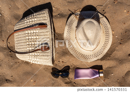 Flat Lay of Beach Essentials Woven Straw Bag Hat Sunglasses and Cream Tube on Sand Flat Lay of Beach Essentials Woven Straw Bag Hat Sunglasses and Cream Tube on Sand 141570331