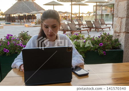 Young Brunette Woman Working on Tablet at Resort Outdoor Cafe Young Brunette Woman Working on Tablet at Resort Outdoor Cafe 141570340