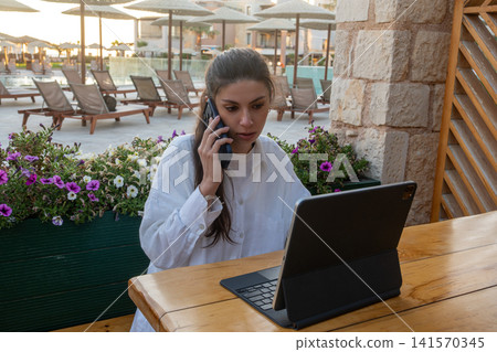 Young Female Entrepreneur Talking on Mobile Phone with Open Tablet Computer at Resort Terrace Young Female Entrepreneur Talking on Mobile Phone with Open Tablet Computer at Resort Terrace 141570345