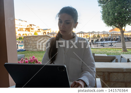 Young Brunette Woman Focusing on Tablet Screen at Resort Outdoor Patio Young Brunette Woman Focusing on Tablet Screen at Resort Outdoor Patio 141570364