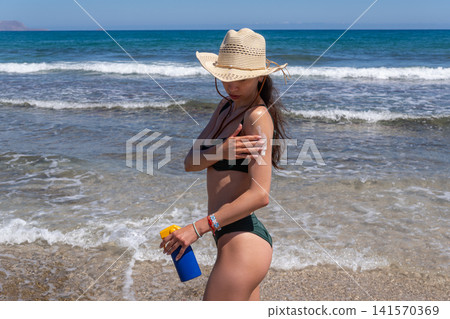 Brunette Woman Applying Sunscreen Lotion on Beach with Sea Waves Background Brunette Woman Applying Sunscreen Lotion on Beach with Sea Waves Background 141570369