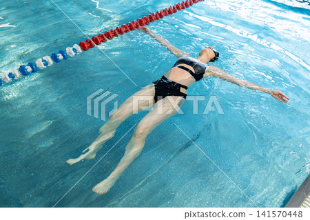 Young woman in black swimsuit swimming in the pool, feet in water, resting near lane divider inside modern swimming facility with wet hair, calm posture and natural light Young woman in black swimsuit swimming in the pool, feet in water, resting near lane divider inside modern swimming facility with wet hair, calm posture and natural light 141570448