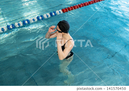 Mature woman adjusting swim goggles in indoor lap pool, wearing swim cap and focused before training, waist-deep in calm water with bright windows and modern wooden ceiling Mature woman adjusting swim goggles in indoor lap pool, wearing swim cap and focused before training, waist-deep in calm water with bright windows and modern wooden ceiling 141570450