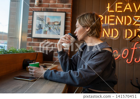 Young woman at cafe counter stirring hot drink beside window with phone, potted plants, casual cutout shoulder outfit, relaxed urban lifestyle mood in bright daylight Young woman at cafe counter stirring hot drink beside window with phone, potted plants, casual cutout shoulder outfit, relaxed urban lifestyle mood in bright daylight 141570451