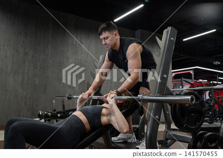 Male trainer spotting female athlete during barbell bench press inside modern gym, focused safety support, strength workout, weight room equipment, determination, and intense training atmosphere Male trainer spotting female athlete during barbell bench press inside modern gym, focused safety support, strength workout, weight room equipment, determination, and intense training atmosphere 141570455