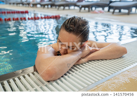 Young person resting beside indoor pool edge with folded arms, wet short hair, calm pensive expression, blue water reflections, tiled deck, and quiet wellness setting Young person resting beside indoor pool edge with folded arms, wet short hair, calm pensive expression, blue water reflections, tiled deck, and quiet wellness setting 141570456