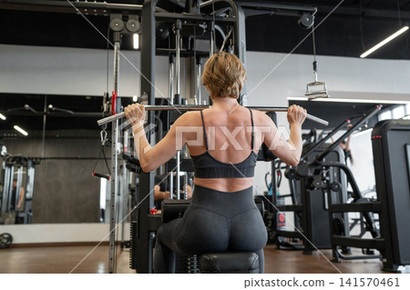 Female athlete performing lat pulldown on cable machine in modern gym from rear view, showing back muscles, focused posture, controlled grip, short hair, sports bra Female athlete performing lat pulldown on cable machine in modern gym from rear view, showing back muscles, focused posture, controlled grip, short hair, sports bra 141570461
