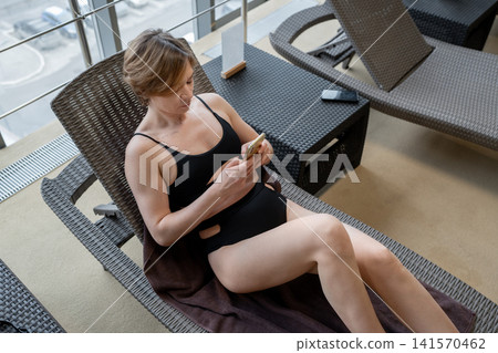 Woman in black swimsuit relaxing on indoor pool lounge chair, using smartphone beside floor-to-ceiling windows and bright city view, with calm natural daylight and modern spa interior 141570462