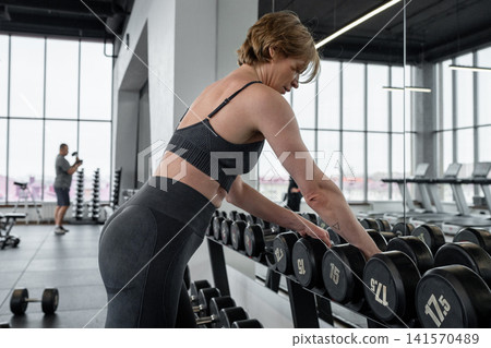 Athletic woman performing one-arm dumbbell row beside rack inside bright modern gym, focused posture and controlled strength training with visible back engagement and core stability Athletic woman performing one-arm dumbbell row beside rack inside bright modern gym, focused posture and controlled strength training with visible back engagement and core stability 141570489