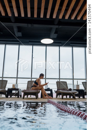 Woman in black swimwear resting on poolside lounger beside indoor pool, hands in wet hair, bright floor to ceiling windows casting backlit silhouette, with woman, swimwear, poolside, lounger detail Woman in black swimwear resting on poolside lounger beside indoor pool, hands in wet hair, bright floor to ceiling windows casting backlit silhouette, with woman, swimwear, poolside, lounger detail 141570494
