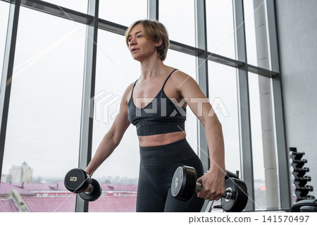 Athletic woman lifting dumbbells in lateral raise exercise inside bright modern gym with floor to ceiling windows and city skyline, focused posture and strong toned arms Athletic woman lifting dumbbells in lateral raise exercise inside bright modern gym with floor to ceiling windows and city skyline, focused posture and strong toned arms 141570497