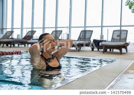 Mature woman adjusting swim goggles in indoor lap pool, wearing swim cap and focused before training, waist-deep in calm water with bright windows and modern wooden ceiling Mature woman adjusting swim goggles in indoor lap pool, wearing swim cap and focused before training, waist-deep in calm water with bright windows and modern wooden ceiling 141570499