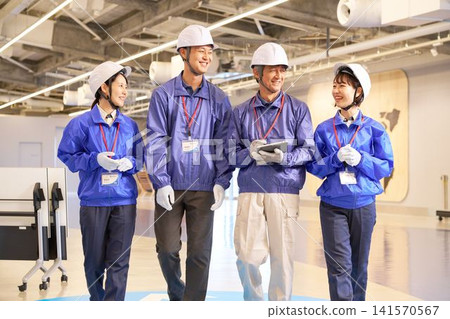 Four workers, two men and one woman, walking with smiles on their faces while wearing helmets. Four workers, two men and one woman, walking with smiles on their faces while wearing helmets. 141570567