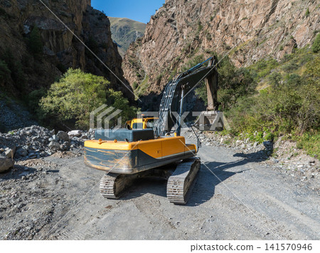 Yellow Excavator on Gravel Road in Mountain Gorge Yellow Excavator on Gravel Road in Mountain Gorge 141570946