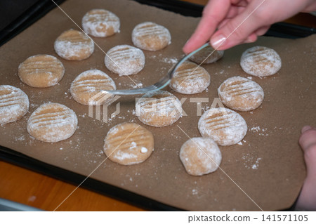 Female hand presses almond cookie dough with fork creating classic cookie pattern on baking tray. Home baking, recipe, handmade dessert, culinary skills, confectionery, preparation 141571105