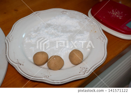 Three almond cookie dough balls rest on powdered sugar before baking on decorative plate in home kitchen. Homemade baking, dessert preparation, recipe, confectionery, cooking, food styling, culinary Three almond cookie dough balls rest on powdered sugar before baking on decorative plate in home kitchen. Homemade baking, dessert preparation, recipe, confectionery, cooking, food styling, culinary 141571122