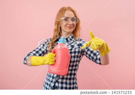 Young woman wearing yellow rubber gloves and safety glasses, holding a pink bottle of cleaning liquid, and pointing down, ready for household chores and demonstrating a product Young woman wearing yellow rubber gloves and safety glasses, holding a pink bottle of cleaning liquid, and pointing down, ready for household chores and demonstrating a product 141571233