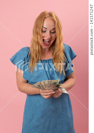 Cheerful young woman in a denim dress holding a fan of dollar banknotes, feeling excited and shocked with a wide open mouth expression on a pink background Cheerful young woman in a denim dress holding a fan of dollar banknotes, feeling excited and shocked with a wide open mouth expression on a pink background 141571247