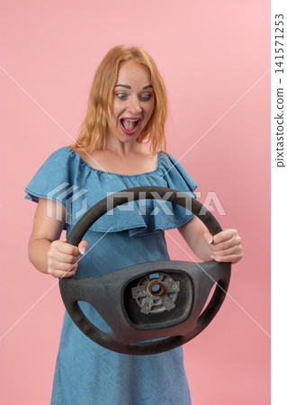 Woman in a blue dress reacting with surprise and shock while holding a damaged. Missing parts automobile steering wheel against a pink background. Expressing concept of unforeseen problems or bad luck Woman in a blue dress reacting with surprise and shock while holding a damaged. Missing parts automobile steering wheel against a pink background. Expressing concept of unforeseen problems or bad luck 141571253