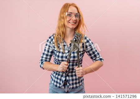 Smiling woman holding a heavy pipe wrench, wearing protective safety glasses and a plaid shirt, standing confidently on a pink background, representing home improvement and female skills Smiling woman holding a heavy pipe wrench, wearing protective safety glasses and a plaid shirt, standing confidently on a pink background, representing home improvement and female skills 141571296