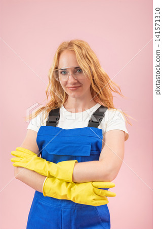 Young woman in protective workwear. Including blue overalls. Yellow gloves. And safety glasses. Standing confidently with crossed arms against a pink background Young woman in protective workwear. Including blue overalls. Yellow gloves. And safety glasses. Standing confidently with crossed arms against a pink background 141571310
