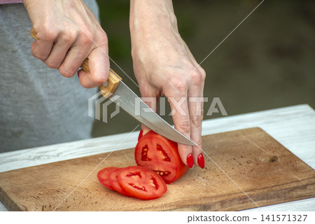 Person's hands holding a knife, slicing a ripe red tomato into thin pieces on a wooden cutting board, preparing ingredients for a healthy meal Person's hands holding a knife, slicing a ripe red tomato into thin pieces on a wooden cutting board, preparing ingredients for a healthy meal 141571327