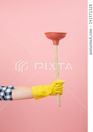 Person's gloved hand extending across a pink background, holding a household plunger, symbolizing cleaning, plumbing, solution, and readiness for tasks Person's gloved hand extending across a pink background, holding a household plunger, symbolizing cleaning, plumbing, solution, and readiness for tasks 141571328