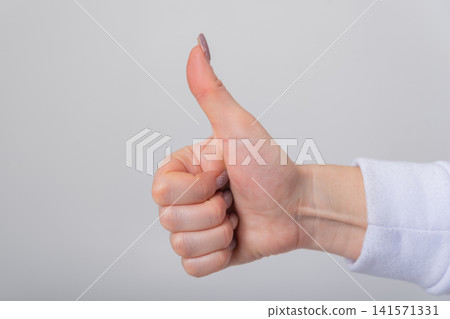 Female hand with polished nails performing a universally recognized thumbs up gesture, symbolizing agreement, success, and positive feedback on a neutral background Female hand with polished nails performing a universally recognized thumbs up gesture, symbolizing agreement, success, and positive feedback on a neutral background 141571331