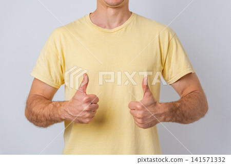 Man in a yellow t shirt presenting two thumbs up, expressing acceptance and agreement, symbolizing approval, success, and positive feedback on a grey background 141571332