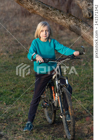 Young blonde boy holding mountain bike handlebars outdoors at golden hour, full length portrait showing active, carefree childhood, cycling adventure and summer freedom Young blonde boy holding mountain bike handlebars outdoors at golden hour, full length portrait showing active, carefree childhood, cycling adventure and summer freedom 141571342