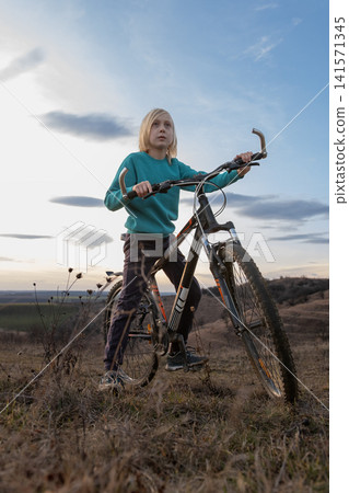 Young boy with blonde hair sitting on a mountain bike on a grassy hill, looking up at the sky, representing concepts of childhood adventure, freedom, and future aspirations Young boy with blonde hair sitting on a mountain bike on a grassy hill, looking up at the sky, representing concepts of childhood adventure, freedom, and future aspirations 141571345