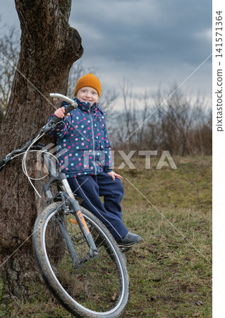 Young child wearing a colorful jacket and hat, smiling while resting with a bicycle against an old tree trunk, enjoying a casual outdoor moment on a cloudy day 141571364