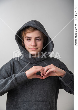 Young person in a grey hooded sweatshirt looks at camera and makes a heart with both hands, expressing love, kindness and gratitude in a casual studio portrait Young person in a grey hooded sweatshirt looks at camera and makes a heart with both hands, expressing love, kindness and gratitude in a casual studio portrait 141571399