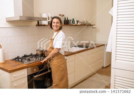 Smiling middle aged woman in apron near stove Smiling middle aged woman in apron near stove 141571662