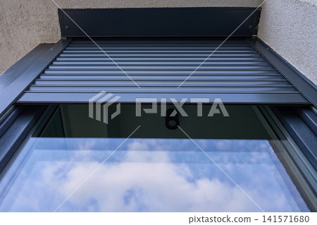 Partially lowered metal roller shutter covers the upper section of an exterior window reflecting sky and clouds Partially lowered metal roller shutter covers the upper section of an exterior window reflecting sky and clouds 141571680