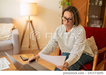 Middle aged female specialist analyzing paper near laptop Middle aged female specialist analyzing paper near laptop 141571695