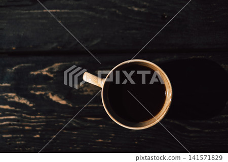 Minimalist top view of a black coffee cup on a dark wooden textured table illuminated by warm natural light. Cozy morning atmosphere with strong shadows, copy space and lifestyle appeal. Wood texture. Minimalist top view of a black coffee cup on a dark wooden textured table illuminated by warm natural light. Cozy morning atmosphere with strong shadows, copy space and lifestyle appeal. Wood texture. 141571829